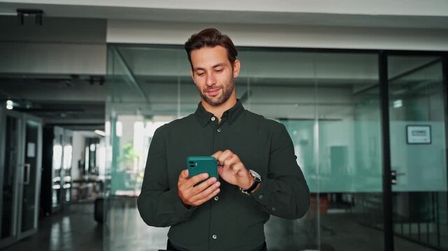 Young European business man manager ceo in green shirt using cell phone mobile app. Smiling Latin Hispanic male businessman holding smartphone in office workplace for trading, reading, working online