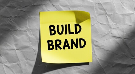 Obraz premium Yellow sticky note with the words Build Brand on a textured grey paper surface with light shadows.