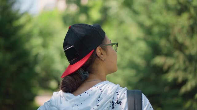 person wearing cap from behind on shaded trail, ponytail tucked through snapback, close framing on hat and dotted hoodie, soft bokeh of evergreen trees, tranquil summer pause and subtle motion