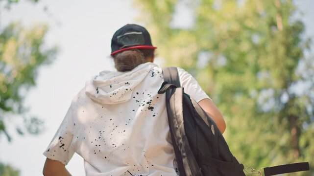 young person walking with backpack outdoors, back view of capwearing youth in hoodie striding toward sunlit forest canopy, dynamic motion and confident pace, textured shirt and shoulder strap