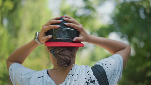 asian young adult adjusting cap backward on shaded path, ponytail tucked under hat, hands to head in motion, readying for walk or hike, casual hoodie and backpack strap details in leafy summer setting