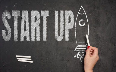 A hand draws the outline of a rocket taking off with white chalk on a black chalkboard, next to which is the large inscription START UP