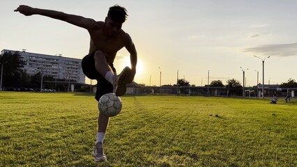 Young man juggling soccer ball on stadium at sunset. Professional footballer kicking ball at green field. Sportsman practicing tricks at meadow with sunlight at background. Freestyle football