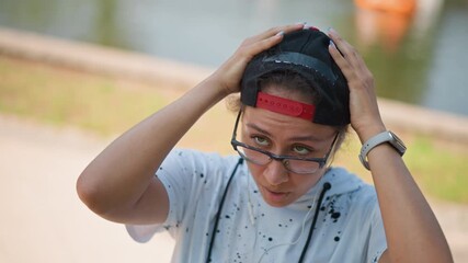 park person adjusting earbuds near lake, fiddling with cord and checking fit, glancing at water and path, cap backward and glasses, sunlight casting soft shadows, casual stroll interrupted