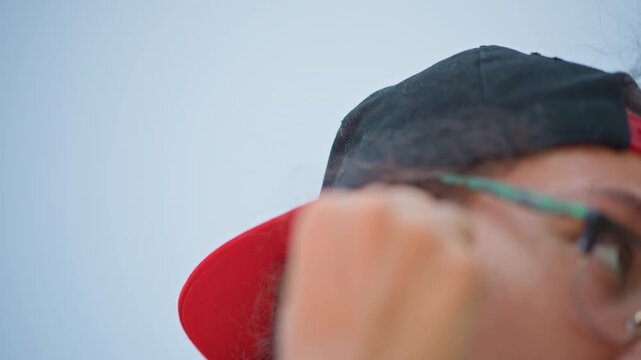 streetside portrait young adult fitting earbuds and glasses under backward cap city sky backdrop, poised pause before moving, wireless audio connection, calm focus and urban routine captured