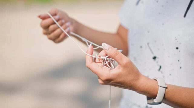 caucasian woman hands untangling earbuds near beach, smartwatch on wrist, white cords twisted, slow patient movements, closeup of fingers solving knot, casual tshirt, sandy blurred background, scenes