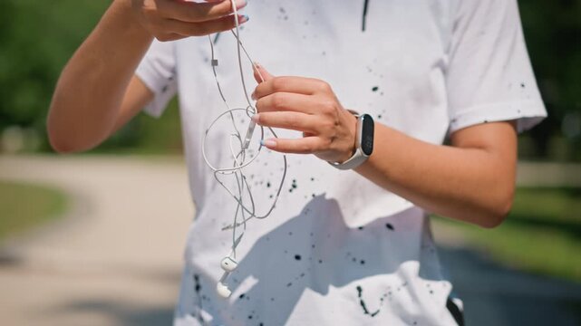 musician untangling earbuds on path, careful hands sorting knotted cable near park walkway, smartwatch visible, focused preparation for rehearsal, natural light and calm atmosphere