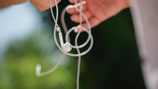 female hands unknotting earbuds in sunlight, garden bokeh backdrop, wrist bracelet and polished nails, gentle breeze, cable loops and slow deliberate motions, outdoor leisure and music preparation