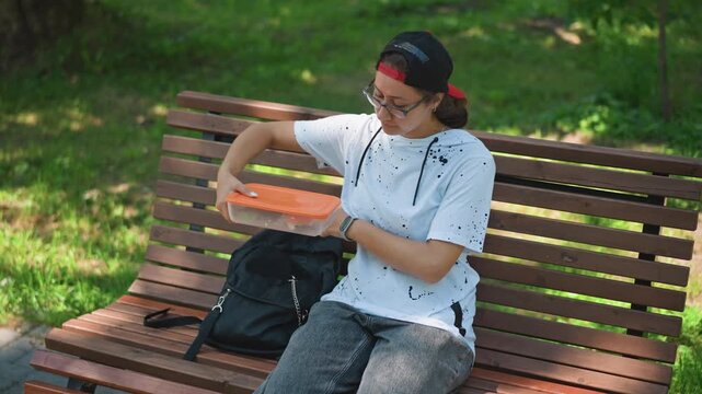 commuter retrieving salad from backpack on bench, black bin nearby, red visor cap, white speckled shirt, orange container open, mixing greens, mindful bite, sunlight and shade, casual routine, outdoor