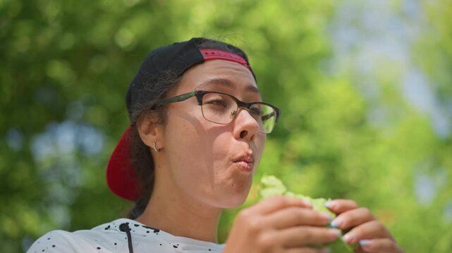 foodie tasting green sandwich under trees, vloggerstyle closeup with glasses and red cap, chewing thoughtfully, park picnic atmosphere with sunlight and candid framing, flavorfocused mood