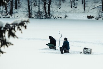 Fishermen fishing on a frozen lake in winter with fishing pole or rod, ice auger and equipment for...