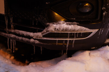 Close-up of a dark car front grille and bumper heavily coated in frost and hanging icicles, illustrating the effects of extreme winter temperatures.