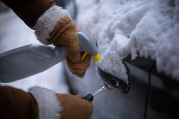 A gloved hand holds a car key while spraying a de-icer liquid into a frozen lock on a snow-covered vehicle to restore access.