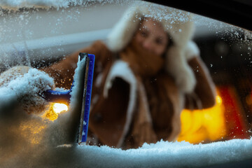 From inside the car, a woman in a fur-lined hood clears a thick layer of snow from the windshield using a blue scraper under city lights at dusk.