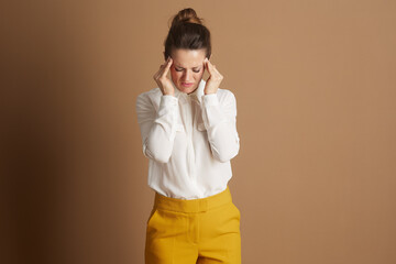 A professional woman in a white shirt and yellow trousers grimaces in pain, holding her temples, suggesting a stress headache or discomfort in a corporate setting.