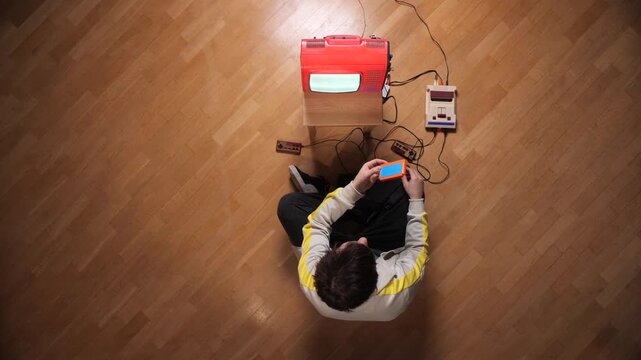 Young man sitting on a wooden floor, inserting a game cartridge into a retro console while connected to an old television, enjoying a nostalgic gaming experience
