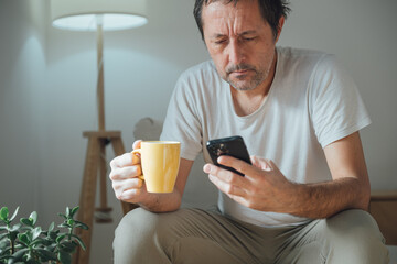 Casual mature man with messy hair drinking morning coffee and scrolling social media on smartphone in cozy living room interior, relaxed home routine lifestyle.
