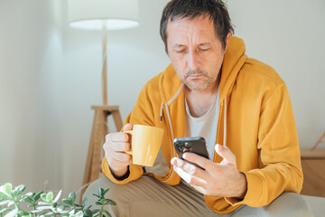 Unshaven adult man enjoying coffee while browsing social media on phone in comfortable home living room, laid-back morning mood.