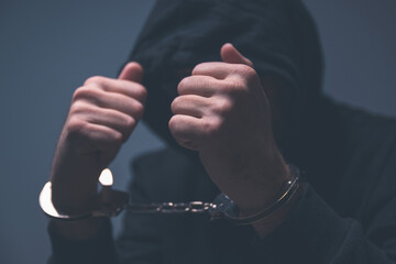 Dark moody image of a hooded offender hiding his face behind tight clenched fists, cuffs reflecting light, conveying anxiety, guilt, and emotional distress.