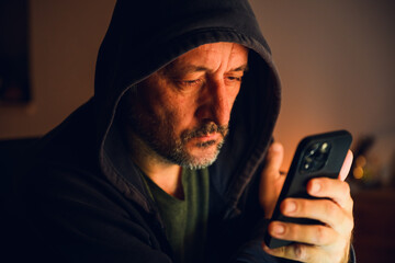Anonymous figure in black hoodie illuminated by warm light, using smartphone in the dark &mdash; mystery and solitude concept.