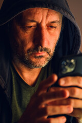 Low key cinematic portrait of man in black hoodie illuminated by soft warm light, focused on smartphone in darkness, symbolizing isolation and digital absorption.