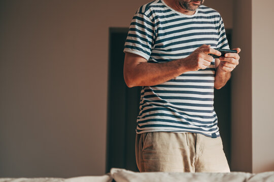 Relaxed middle-aged man in striped t-shirt and beige shorts stands casually, typing intently on his black smartphone with both hands.