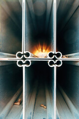 Stacked metallic steel construction profiles forming a cross pattern, with a dramatic golden light burst glowing from the center and dried leaves at the bottom.