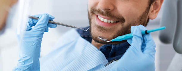 Closeup of dental clinic patient smile and dentist hands holding tools, unrecognizable cheerful bearded man attending dentistry, panorama. Banner for modern dental clinic advertisement
