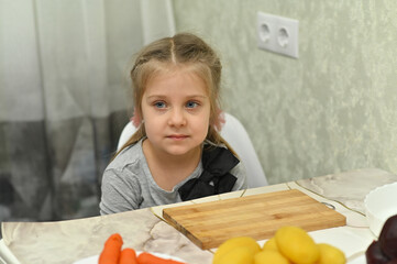 Little girl sits near table with carrot and potato, kitchen background, cooking process, child lifestyle, concept of healthy nutrition. Girl resting near cutting board with vegetables.