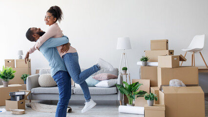 Glad millennial african american husband hugs and lifts his wife in new apartment around cardboard boxes and things, celebrate moving and buying apartment. Moving, relocation together, free space