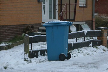blue plastic recycling bin for paper waste stands at the roadside, covered in snow, ready for collection