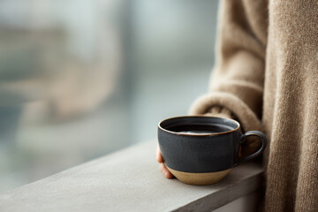 Individual holds a warm cup of tea near a window, surrounded by soft natural light, evoking a peaceful and cozy mood in a minimal setting