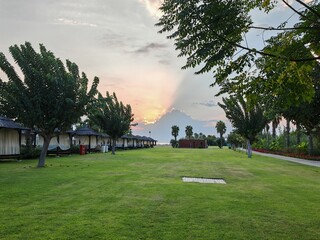 Wooden gazebos with thatched roofs and curtains stand along the sea beach. Nearby is a mowed lawn,  palms and other trees. The autumn sunset sun shines through the clouds and paints the sky