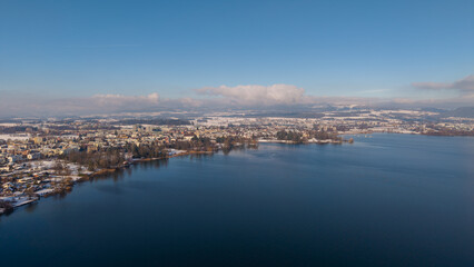 Fototapeta premium Aerial drone view along the shore of Hünenberg See shows snow-covered homes and residential blocks beside the deep blue Lake Zug under a clear, sunny winter sky.