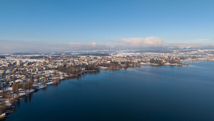 Fototapeta premium Aerial drone view along the shore of Hünenberg See shows snow-covered homes and residential blocks beside the deep blue Lake Zug under a clear, sunny winter sky.