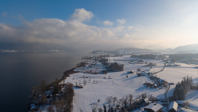 Aerial view of the snow-covered western shore of Lake Zug toward Risch, with Mount Rigi partly wrapped in clouds, creating a calm, atmospheric winter landscape. - Powered by Adobe