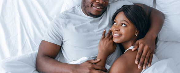 Two people lie together in bed, smiling and looking at each other. They appear happy and...