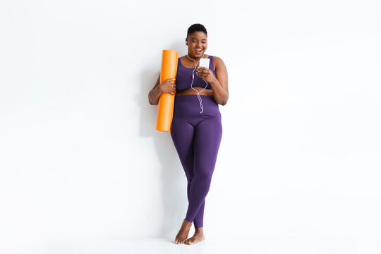 A woman is standing by a white wall with an orange yoga mat. She is looking at her phone and listening to music, wearing workout clothes. It is bright and she appears to be in a fitness studio.
