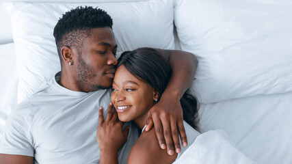 Two people relax in bed, wrapped in each other's arms. They smile at each other, enjoying a moment...