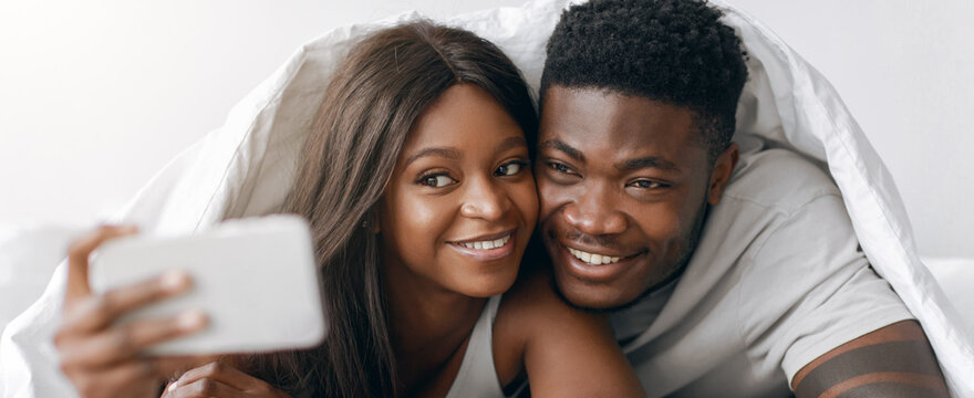 A couple enjoys their time together while lying under a blanket in their bedroom. They smile as they take a selfie with a phone. This moment captures their joy and connection. - Powered by Adobe