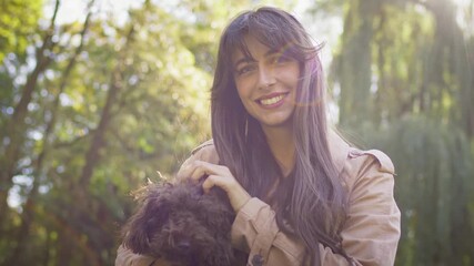 Cheerful Caucasian woman with long hair smiling at camera while holding small brown dog outdoors. Female enjoying sunny day in park surrounded by soft foliage and golden afternoon light.