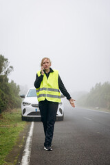 Female roadside assistance technician in reflective vest is communicating on phone while standing beside a white vehicle on a foggy road, showcasing emergency service and support