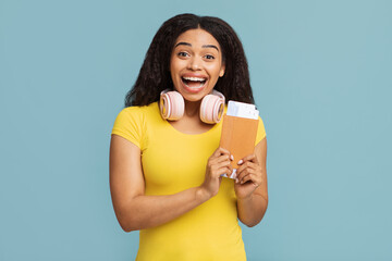 Cheap flights concept. Overjoyed black woman holding passport and boarding pass tickets, standing isolated on blue background. Excited lady with headphones on neck going on vacation after lockdown