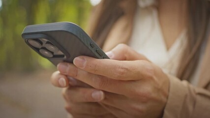 Close up of female hands holding smartphone outdoors typing on screen. Woman wearing beige coat using mobile device while standing in park with blurred trees. Remote communication concept.