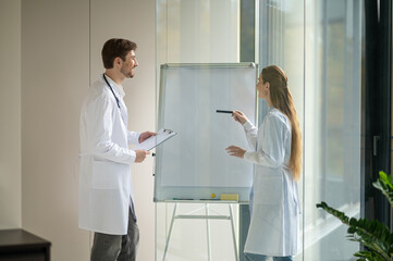 Obraz premium Female doctor standing near the flipchart and presenting something to a new doctor