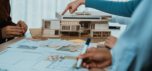 A group of architects engages in an intense discussion about building design, showcasing a scale model and multiple blueprints on a table in a bright workspace. SACTR
