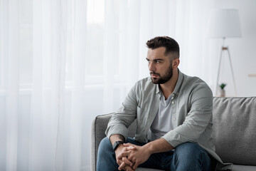 Thoughtful serious bearded millennial male sit on sofa alone at home, lost in thoughts, thinking...