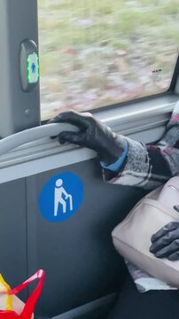 gloved hand on bus panel, unspecified passenger resting by window near priority seating symbol, red shopping bag at feet, motion blurred