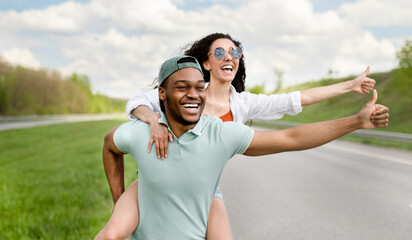 Multiracial couple hitchhiking on road, stopping car with thumb up gesture, traveling by autostop during summer vacation, panorama. Black man giving his girlfriend piggyback ride on roadside