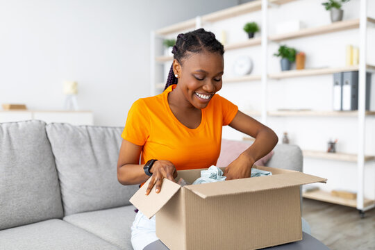 Online delivery service. Cheerful African American woman unpacking parcel box with purchase at home. Happy female customer satisfied with ordered goods, looking at new clothes, shopping on web